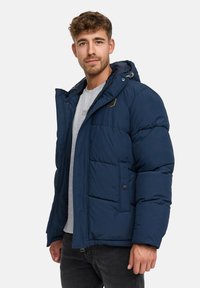 Młody mężczyzna ubrany w granatową ocieplanetą kurtkę z kapturem, pod którą ma na sobie jasnoszary T-shirt, stoi na tle jednolitego białego tła.