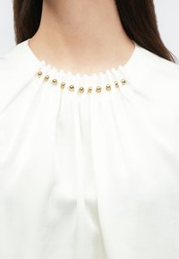 Hvid top med indsamlet stof ved halsudskæringen med guldfarvede accenter og plisseringer, der skaber en tekstureret visuel effekt.