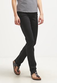 Pantaloni neri slim-fit con un taglio classico, dotati di tasche anteriori e una trama liscia, abbinati a sandali neri piatti su uno sfondo neutro.
