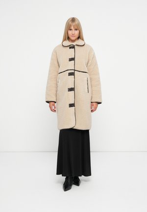 Beige shaggy coat with a rounded collar, black button closures, and black trim, worn over a long black skirt and heels.