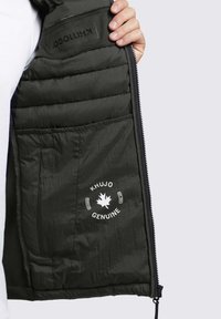 Grüne Puffer-Weste mit gestepptem Innenfutter, schwarzem Reißverschluss und einem weißen Logo mit einem Ahornblatt und dem Text "KHUJO GENUINE".
