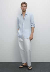 Chemise en lin bleu clair à manches longues, pantalon blanc ample et tongs noires. Design épuré avec boutons et poignets retroussés.