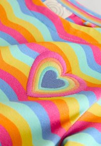 Közelkép egy szivárvány csíkos anyagról, közepén hímzett, többszínű szív alakú folttal.