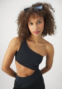 Svart bikinioverdel med én skulder og utskjæringsdesign, mykt stoff, glatt tekstur. Modellen har krusete hår og bruker solbriller; minimale tilbehør.