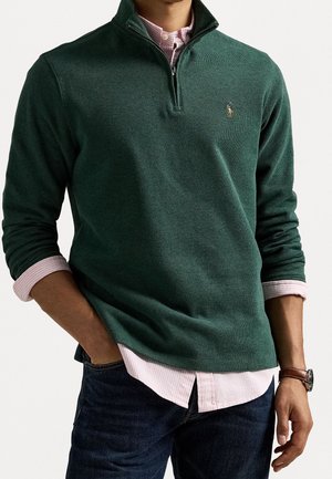 Uomo che indossa un maglione verde scuro con cerniera a quarto di zip sopra una camicia rosa chiaro con bottoni, jeans scuri e orologio da polso marrone, mani rilassate lungo i fianchi.
