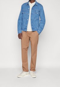 Man wearing light blue denim jacket, white shirt, tan chinos with folded cuffs, and off-white sneakers with white stripes against a plain background.