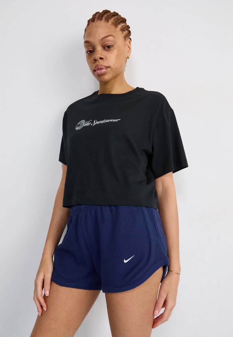 T-shirt nera corta con testo del logo bianco, abbinata a pantaloni corti a costine blu scuro con il logo Nike bianco. Il materiale sembra morbido e traspirante.