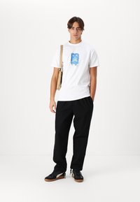 Camiseta blanca con diseño gráfico azul, pantalones negros de corte holgado y zapatillas negras con suelas de goma. El modelo sostiene una bolsa beige.