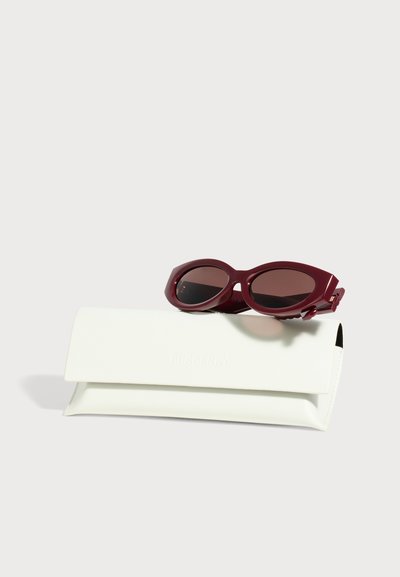 Des lunettes de soleil ovales bordeaux aux verres foncés reposent sur un étui blanc de marque, sur un fond clair.