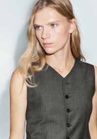 Szaro-sukienka bez rękawów, uszyta z teksturowanej tkaniny, z dekoltem w kształcie litery V i pięcioma czarnymi guzikami z przodu, prezentująca dopasowany krój.