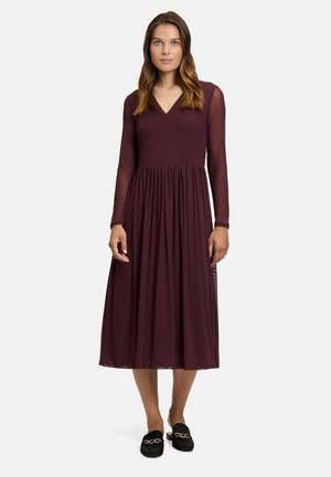 Weinrotes Kleid mit langen transparenten Ärmeln, V-Ausschnitt, elastischem Taillenbund und fließendem knielangen Rock. Kombiniert mit schwarzen Slippers mit goldenen Beschlägen.