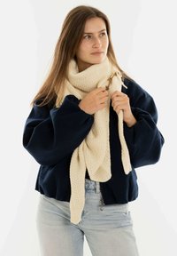 Szydełkowany kremowy szalik z teksturowanym wzorem, luźno przewieszony. Noszony z granatowym, dużym swetrem, podkreślającym swobodny krój i casualowy styl.