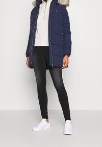 Chaqueta de puff navy con capucha de piel sintética, cierre con cremallera y diseño acolchado. Combinada con leggings oscuros y zapatillas blancas con detalles en amarillo.