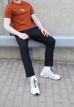 Osoba w pomarańczowej koszulce, czarnych spodniach, białych skarpetkach i białych trampkach siedzi na szarej metalowej ławce przy betonie.