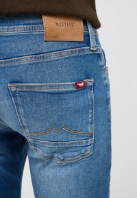 Niebieskie dżinsy z denimu z skórzaną metką oznaczoną "MUSTANG". Ma czerwoną etykietę na tylnej kieszeni oraz kontrastowe detale szwów.