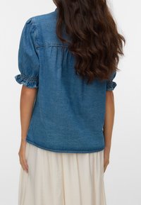 Femme aux longs cheveux bruns ondulés portant un haut en jean bleu à manches courtes avec des manches à volants et une jupe blanche, vue de dos.