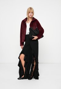 Bordeaux nepsjaal jasje, witte top, zwarte rok met ruches en split, en zwarte flats. Model houdt een zwarte tas vast, minimalistische achtergrond.