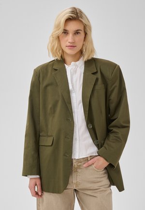 Femme blonde portant un blazer vert olive oversize, une chemise blanche boutonnée et un pantalon beige, une main dans la poche, sur fond uni.