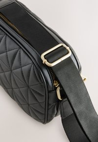 Sort quiltet håndtaske med rektangulært design, gulddetaljer og en glat satinrem med justerbar længde. Lynlåslukning inkluderet.