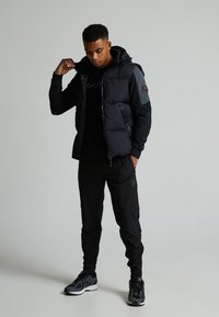 Μαύρο επενδυμένο μπουφάν με κουκούλα, κλείσιμο με φερμουάρ και λογότυπο στο στήθος, συνδυασμένο με μαύρες tapered παντελόνες και αθλητικά μαύρα παπούτσια.