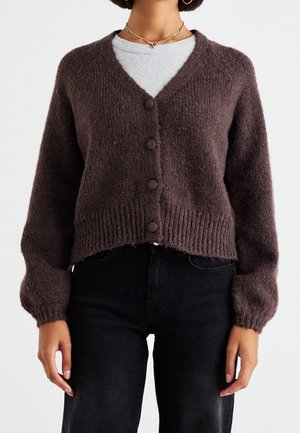 Mujer con un cárdigan marrón oscuro con botones y textura suave, sobre una camiseta gris clara y pantalones negros, recortada a la altura del cuello y la cintura.