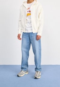 Felpa con cappuccio bianca con cerniera, t-shirt con grafica, jeans larghi azzurro chiaro e sneakers grigie con dettagli in mesh e gomma. Sfondo semplice.