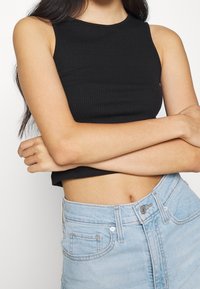 Kobieta z długimi ciemnymi włosami, ubrana w czarny bezrękawnik i jasnoniebieskie dżinsy z wysokim stanem, z założonymi na siebie rękami na brzuchu.