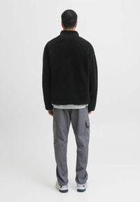 Chaqueta de forro polar negra con cuello alto, tejido texturizado y mangas raglán, llevada sobre una camisa gris claro y combinada con pantalones cargo grises.
