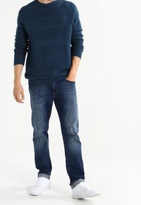 Blaues geripptes Strickpullover mit Rundhalsausschnitt, kombiniert mit ausgewaschenen blauen Jeans und weißen Sportschuhen.