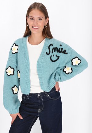 Světle modrý pletený kardigan s oversized rukávy, černými květinovými aplikacemi a slovem "Smile" v černém písmu na přední straně.