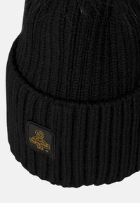 Berretto in maglia nera con una texture a costine e una parte superiore rotonda. Presenta un'etichetta quadrata nera con dettagli in giallo sul logo nella parte anteriore.