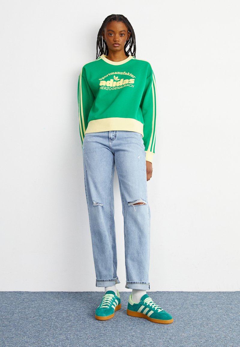 Felpa verde con accenti crema e testo "adidas", abbinata a jeans strappati azzurri chiari e sneakers verdi con strisce bianche.