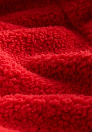 Roter Plüschstoff mit einer flauschigen, strukturierten Oberfläche, die einen tiefen Flor und ein weiches, behagliches Aussehen aufweist, mit sanften Wellen durchzogen.
