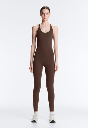 Žena stojaca čelom vpred, oblečená v hnedý bezrukávový overal celotelovej dĺžky s béžovým lemovaním a bielymi teniskami na bielom pozadí.