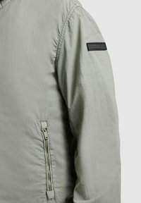 Svetlosivý rukáv bundy a bok s vertikálnym vreckom na zips a čiernou obdĺžnikovou nášivkou na hornej časti ramena.