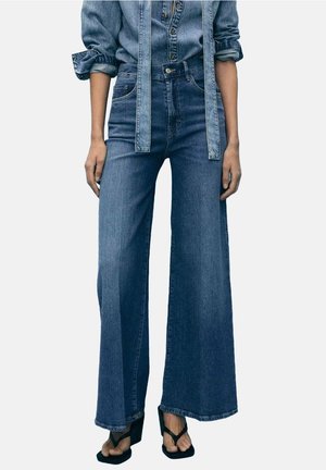 Kobieta ubrana w niebieskie dżinsy z wysokim stanem i szerokimi nogawkami, zestawione z dżinsową koszulą, czarnymi sandałami na obcasie oraz długimi dżinsowymi szelkami.