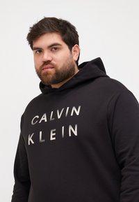 Čierna mikina s kapucňou vyrobená z mäkkej tkaniny, s veľkým metalickým textom "CALVIN KLEIN" na prednej strane a vo voľnom strihu.