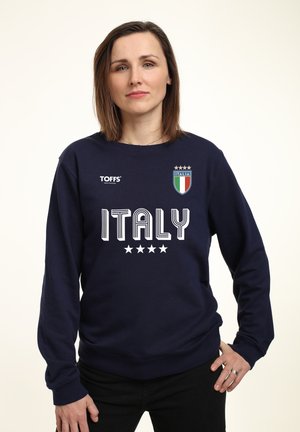 Kvinde med langt, brunt hår iført en marineblå sweatshirt med Italien-logo, teksten "ITALY" og fire stjerner, stående med den ene hånd på hoften.