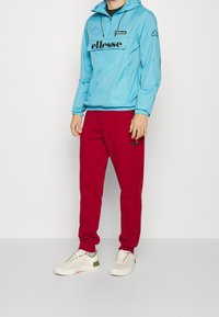 Rompevientos azul claro con capucha y logo negro, combinado con pantalones deportivos rojos; el modelo lleva zapatillas blancas con acentos verdes.