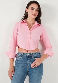 Light pink cropped blouse with a collared neckline, button front, and rolled-up long sleeves, paired with blue high-waisted jeans.
