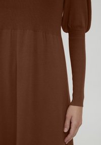 Bruine gebreide jurk met een aansluitend lijfje, pofmouwen en een geribbelde textuur bij de manchetten. Glad materiaal vloeit over in een uitlopende rok.