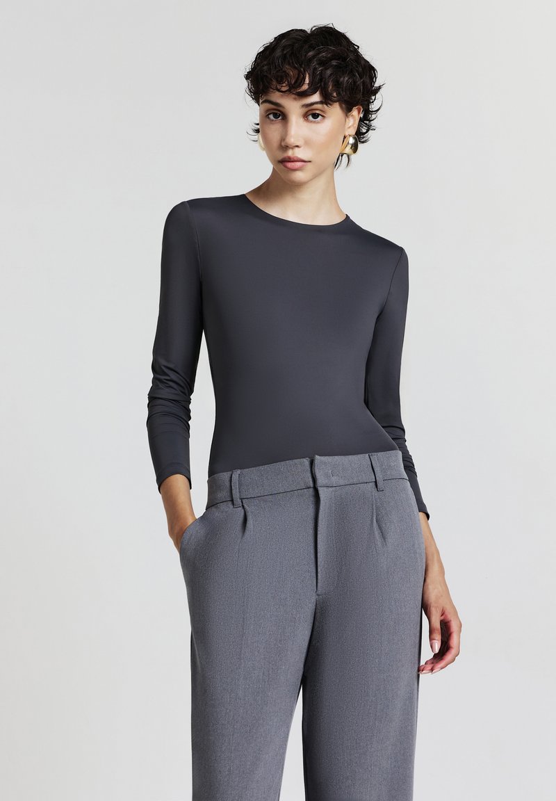 Body grigio a maniche lunghe realizzato in materiale liscio, abbinato a pantaloni grigi su misura con pieghe frontali e tasche oblique.