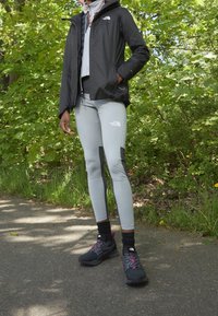 Černá voděodolná bunda, šedá zateplená střední vrstva, šedé legíny s černými akcenty a černé sportovní boty s růžovými detaily na cestě.