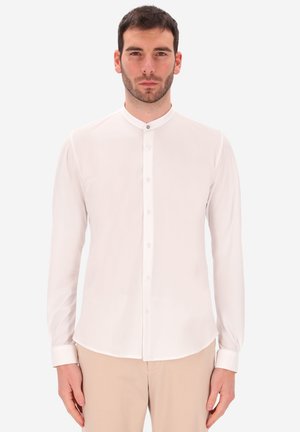 Uomo in piedi che indossa una camicia bianca con colletto alla mandarino e pantaloni beige davanti a uno sfondo chiaro e semplice.