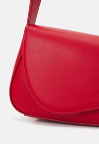 Rød faux læder håndtaske med et buet design, glat tekstur og klaplukning. Har kontrastsyninger langs kanterne.
