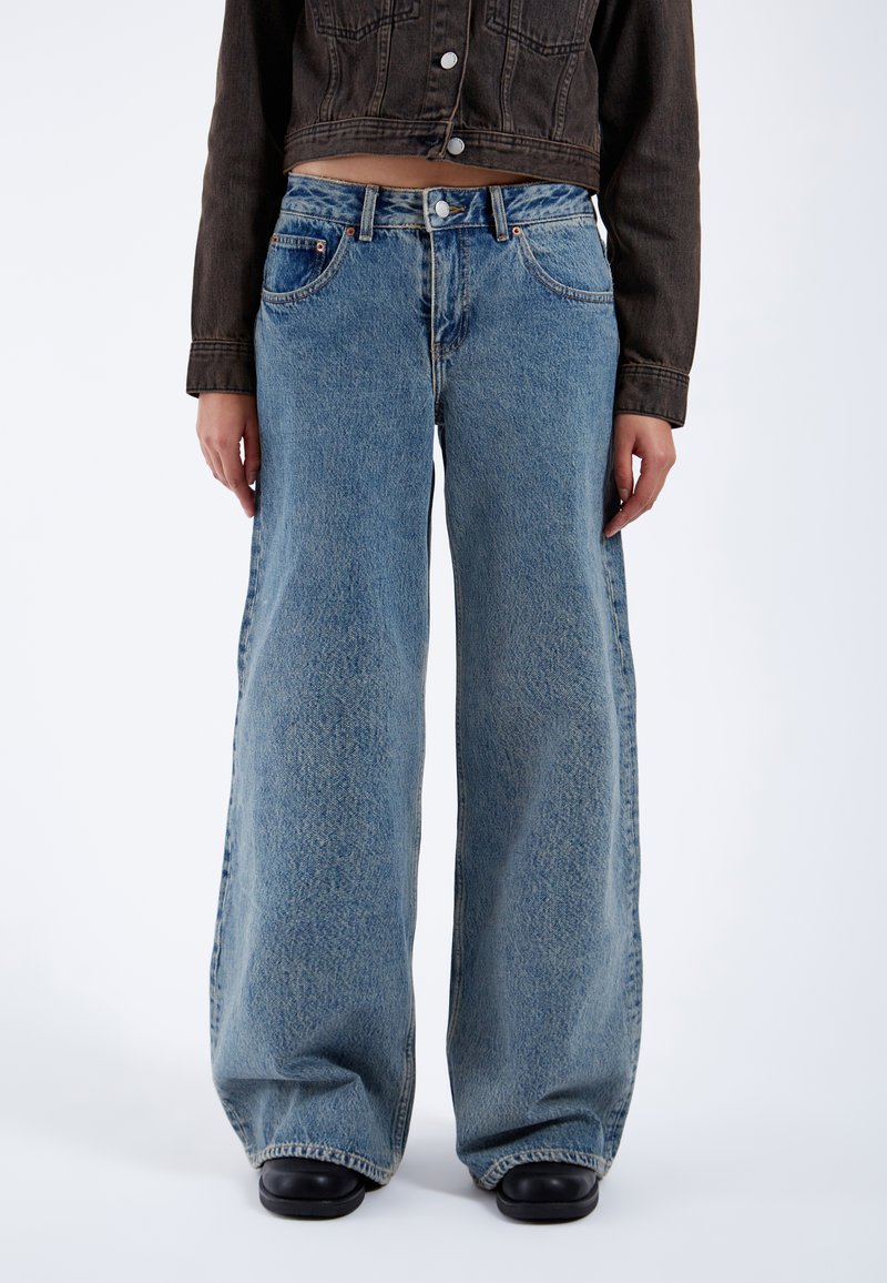 Persona che indossa jeans blu a gamba larga con vita alta, scarpe nere e una giacca corta di denim scuro su uno sfondo semplice.