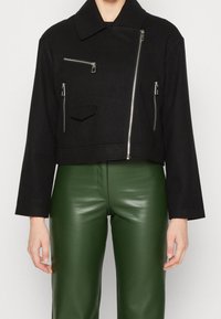 Veste noire courte avec un large col et plusieurs poches zippées. Fabriquée à partir d'un tissu texturé, associée à un pantalon en cuir vert.