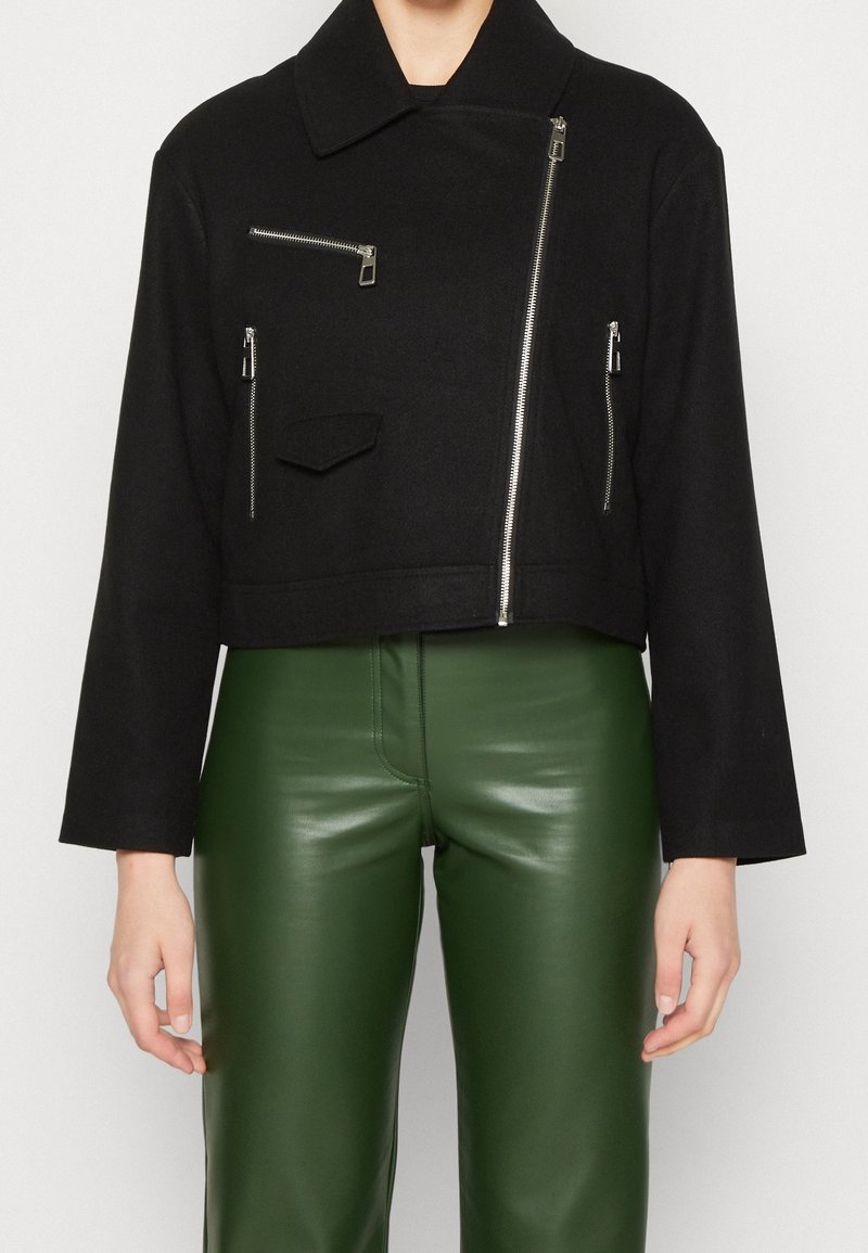 Veste noire courte avec un large col et plusieurs poches zippées. Fabriquée à partir d'un tissu texturé, associée à un pantalon en cuir vert.
