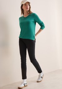 Türkisfarbener Pullover mit einem runden Ausschnitt, kombiniert mit schwarzen, schmal geschnittenen Hosen. Weiße Sneaker mit schwarzen Akzenten. Heller Hintergrund.
