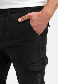 Black cotton cargo pants with a relaxed fit, featuring side pockets and a drawstring waist. Textured fabric and visible stitching details.